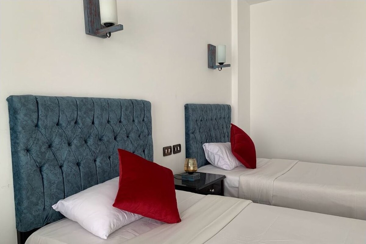 Two beds with blue headboards, white bedding, and red pillows. There are wall-mounted lamps and a small table with a lamp.