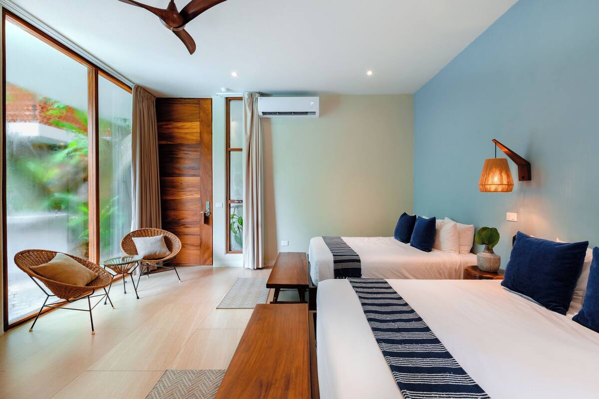 Una habitación de hotel moderna con dos camas, un área de descanso con sillas de mimbre, un banco de madera y un amplio ventanal con vistas a la vegetación.