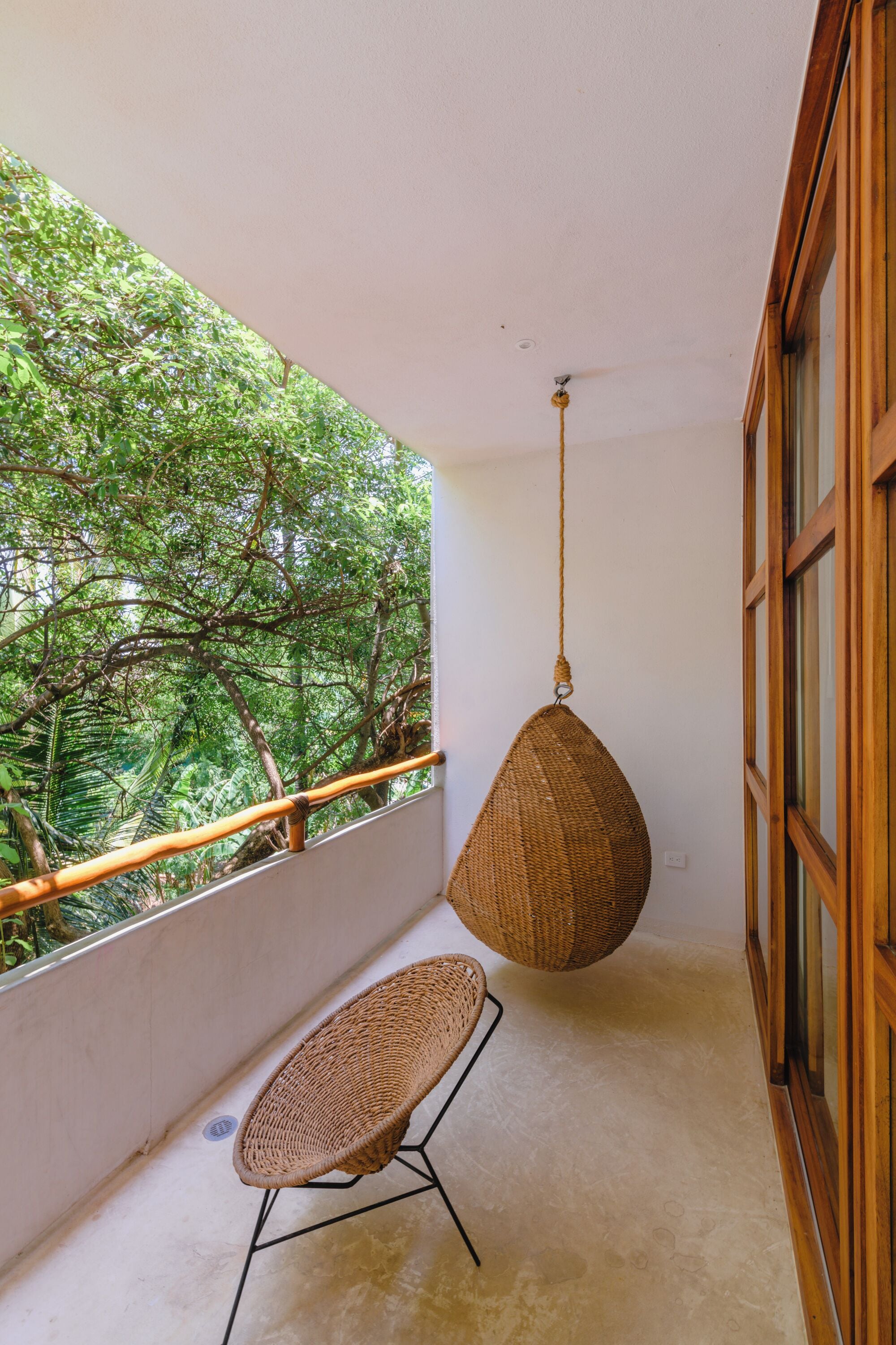Un balcón con una silla de mimbre, una cesta colgante y vistas de vegetación exuberante.