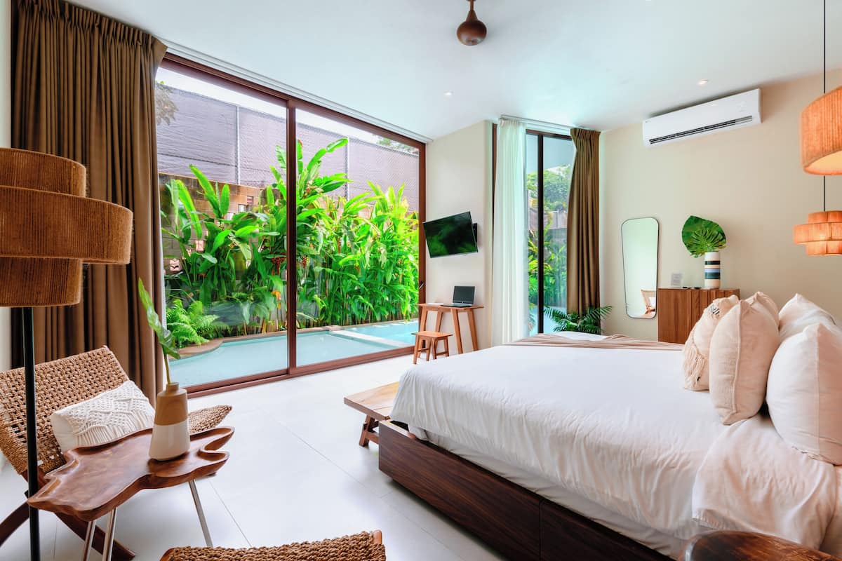 Un dormitorio moderno con una cama grande, un escritorio y vistas a una piscina y follaje.