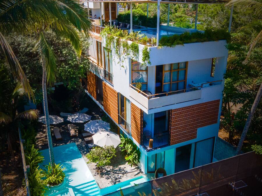 Un edifici modern de diversos pisos amb un jardí al terrat, una piscina i vegetació abundant que envolta la propietat.