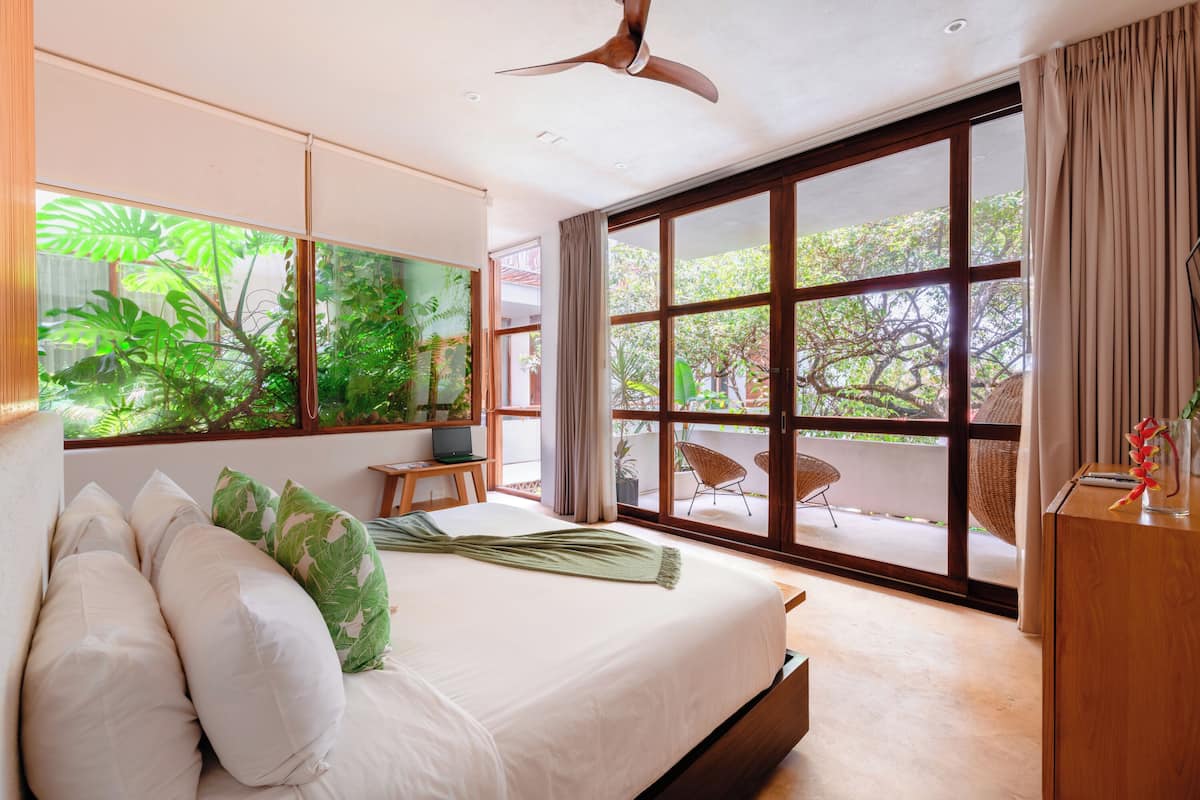 Un dormitorio moderno con una cama grande, un ventilador de techo, un televisor y vistas al exterior a través de amplias ventanas.