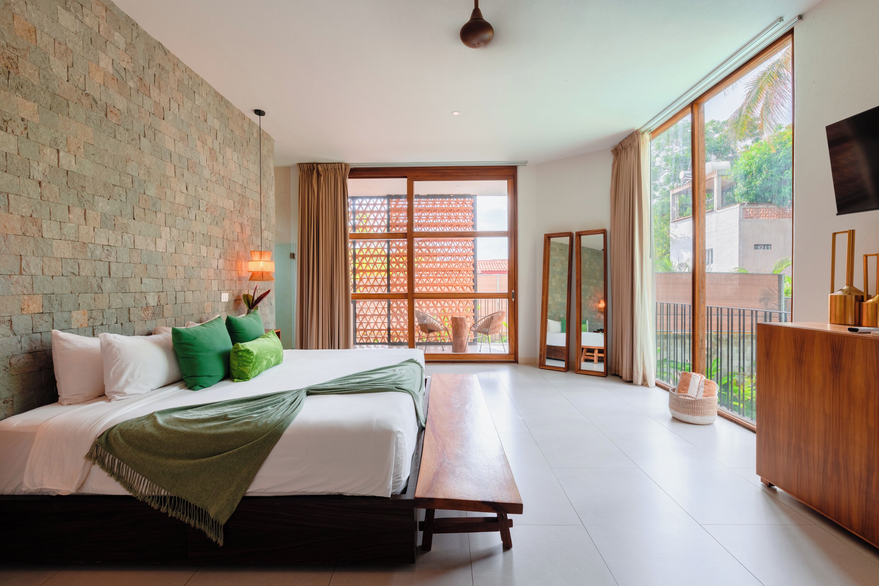 Un dormitorio con una cama grande, un banco de madera, un televisor y un balcón con vistas a la vegetación.