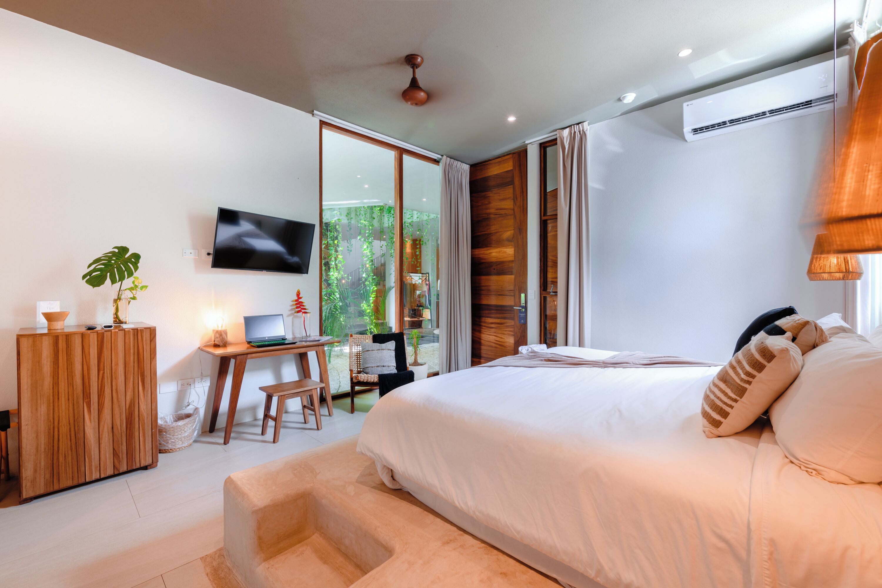 A modern bedroom with a large bed, a desk with a laptop, a television, and a sliding glass door leading to a balcony with greenery.