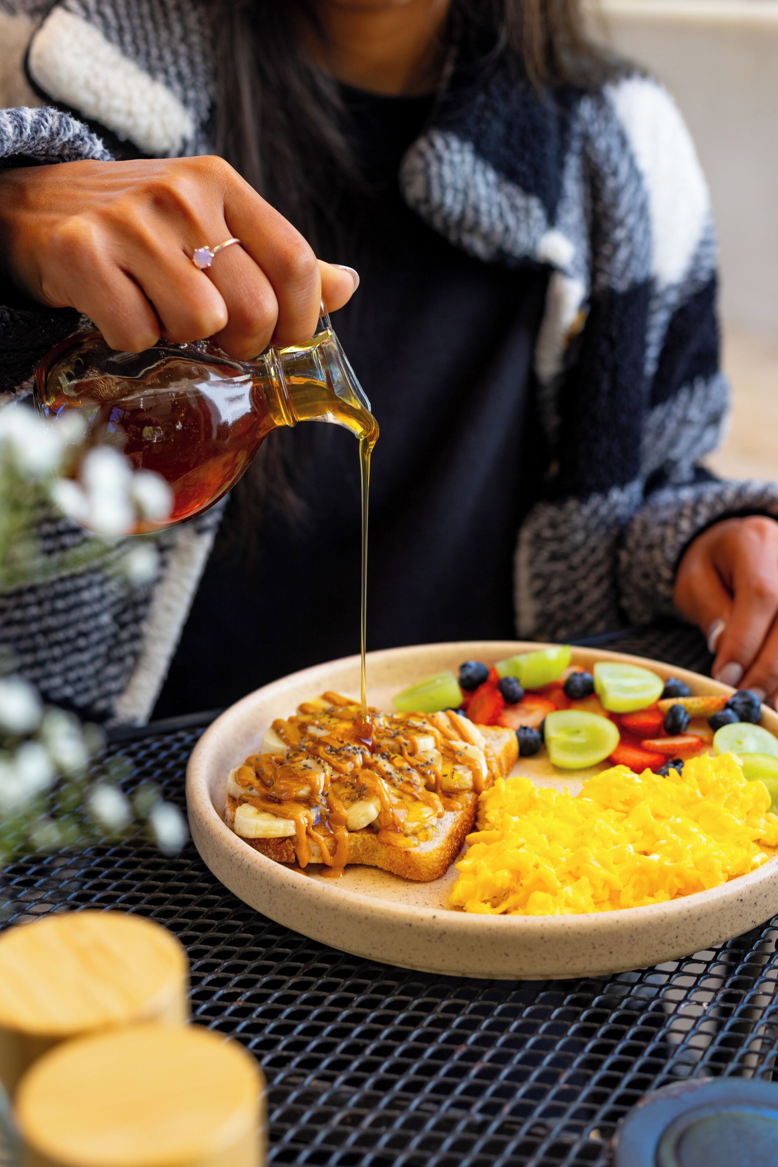 A person pouring syrup over a breakfast plate with toast, fruit, and scrambled eggs.
