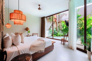 A modern bedroom with a large bed, a desk, and a view of a swimming pool and greenery.
