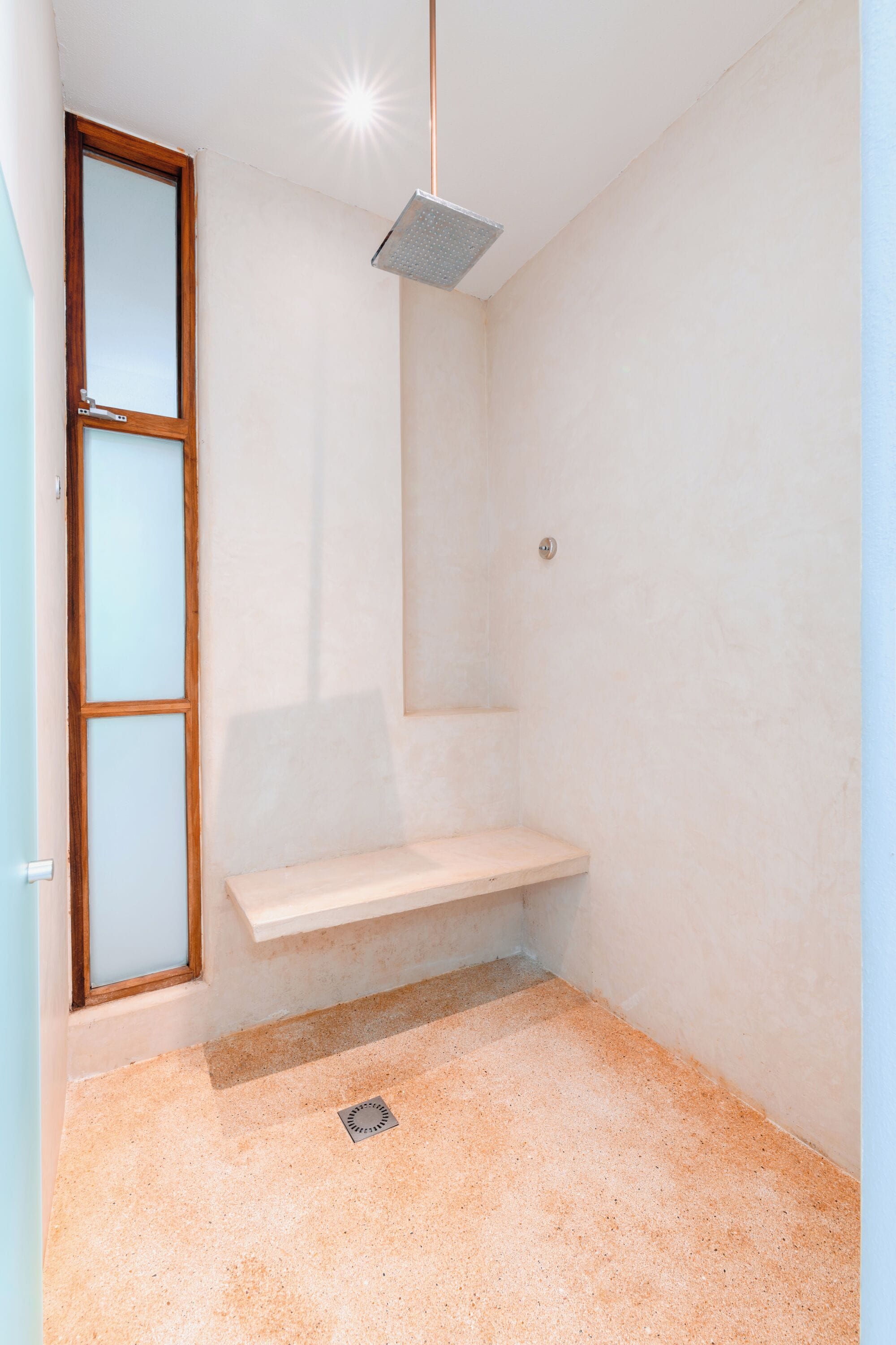 Un área de ducha pequeña y cerrada, con un banco, una ventana y un cabezal de ducha.