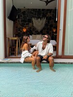 A couple sitting by a pool with a stone wall and a bed in the background.