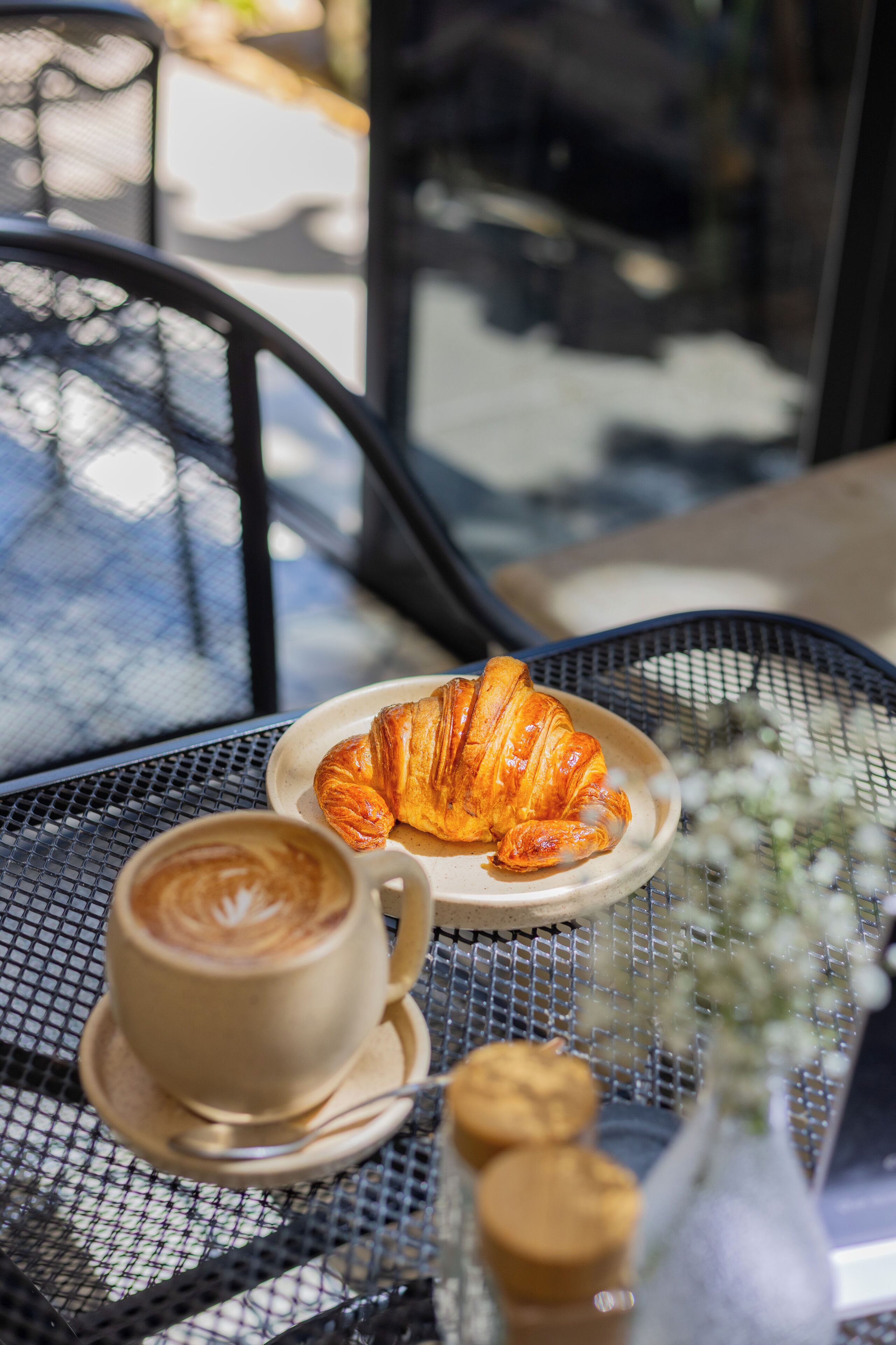 A table with a croissant, a cup of coffee, and a small vase with flowers.