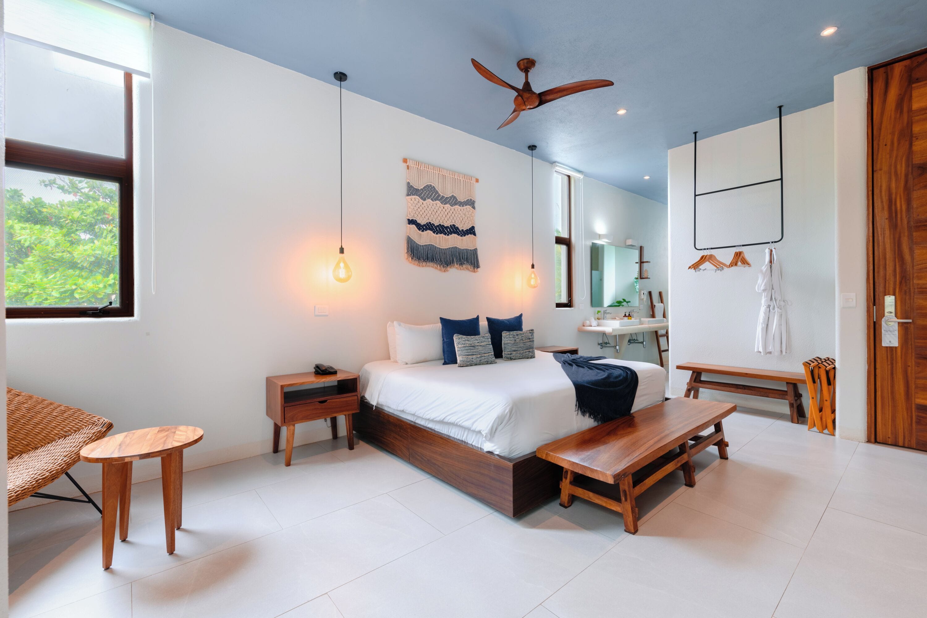 Quarto moderno com cama, mesas de cabeceira, um banco de madeira, ventilador de teto e janela com vista para vegetação.