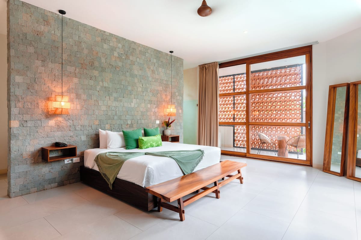 Un dormitorio con pared de piedra, una cama con almohadas verdes, un banco de madera y un amplio ventanal corredero que da a un espacio exterior.