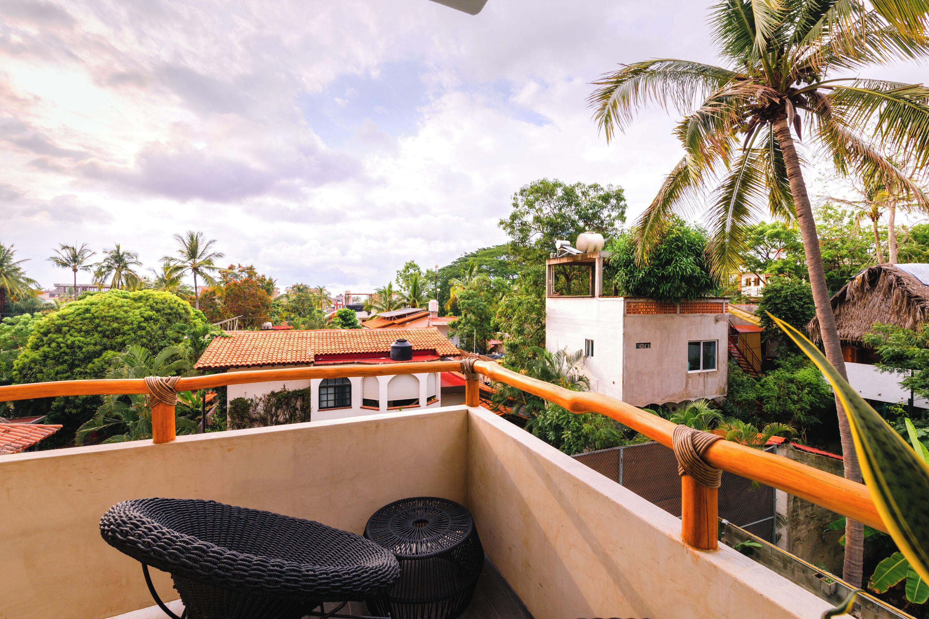 A balcony with a wicker chair and a small table, overlooking a residential area with palm trees and houses.