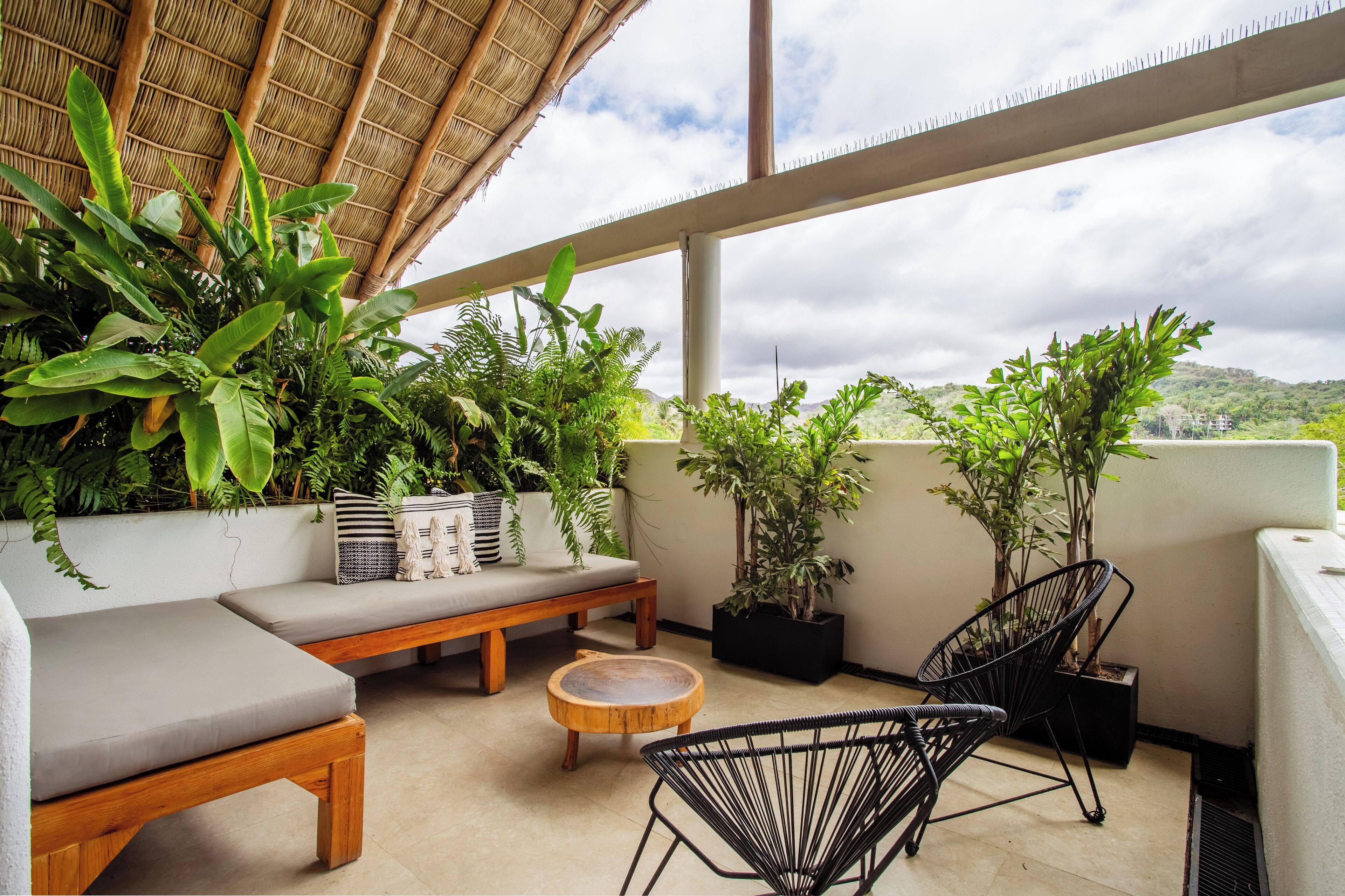 A balcony with a wooden bench, a round wooden table, and two black wire chairs. There are several potted plants and a large leafy plant in the background.