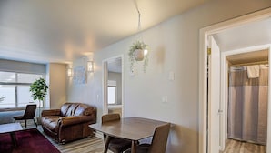 Un salon moderne avec un canapé en cuir marron, une table à manger en bois et des chaises. On y trouve une plante suspendue, un tapis rouge et une fenêtre avec des stores.