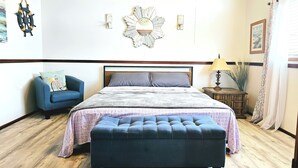 A bedroom with a bed, a blue armchair, a wooden nightstand, a lamp, and a wall-mounted mirror. - CAPTAIN'S CAPTAIN-Waterfront House (Port Arthur)