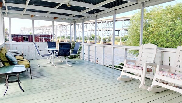 A covered outdoor seating area with tables and chairs, overlooking a body of water and a bridge.