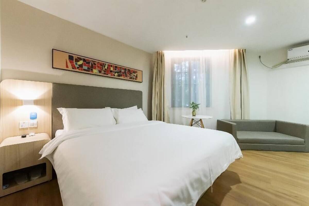 A hotel room with a bed, bedside lamp, a small table with a plant, a grey sofa, and a window with curtains.