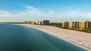 A coastal area with a sandy beach, a clear blue sea, and high-rise buildings in the background. - ⭐ NEW! Starfish Wishes | Saltwater Pool & Spa • Walk to Beach • Sleeps 9 (Naples)