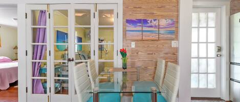 A dining area with a glass table and chairs, a wooden wall with a beach painting, and a door with frosted glass panels.
