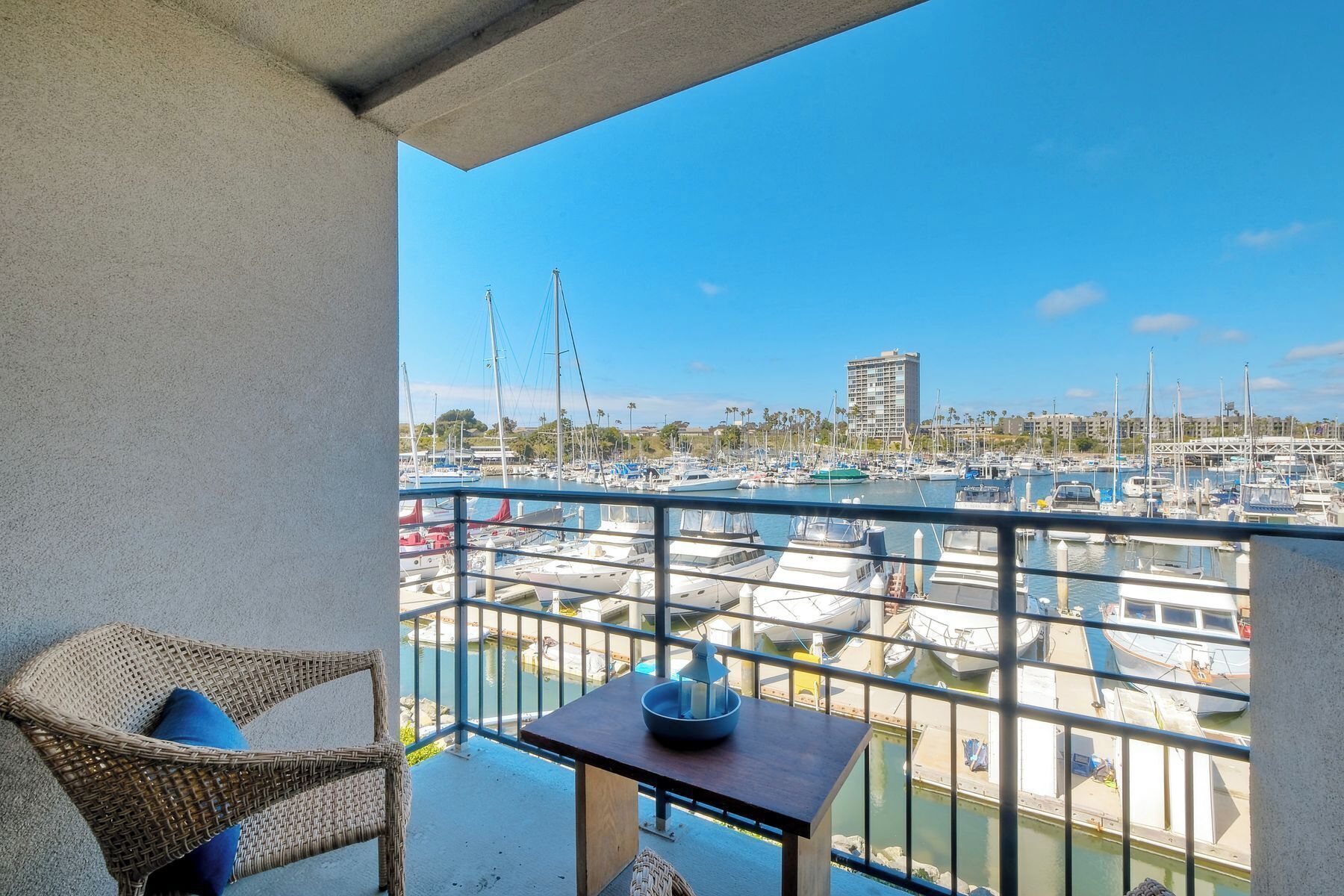 A balcony with wicker furniture and a small table, overlooking a marina with various boats and a distant building.