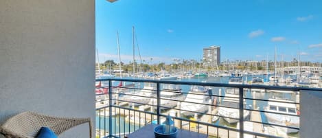 A balcony with wicker furniture and a small table, overlooking a marina with various boats and a distant building.