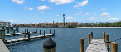 A waterfront hotel with a dock extending into calm water, surrounded by a clear blue sky and distant palm trees.