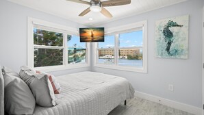 Un dormitorio con cama, ventilador de techo, televisor y ventanas amplias con vistas a edificios y agua.