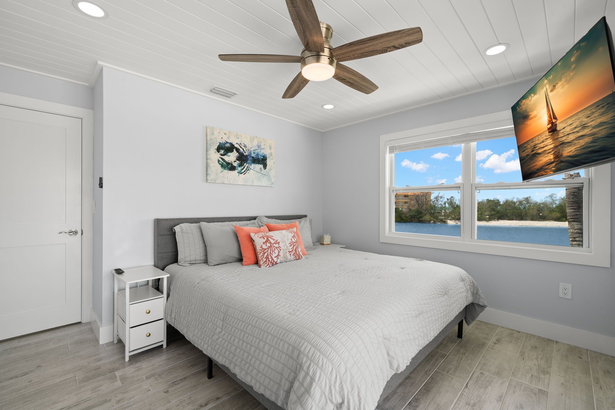 Une chambre à coucher comprenant un lit, une table de chevet, un ventilateur de plafond, un tableau et une fenêtre donnant sur la plage.