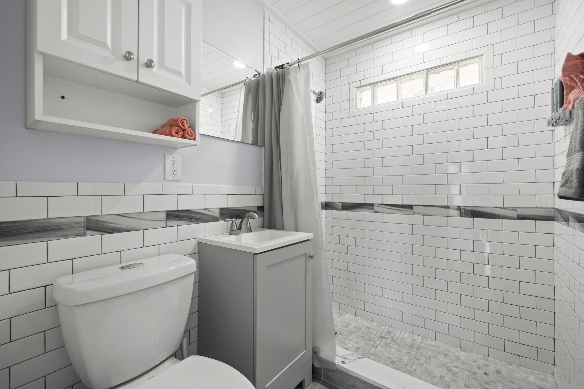 Une salle de bain moderne avec des carreaux de céramique blancs rectangulaires, des toilettes, un lavabo et un espace douche.