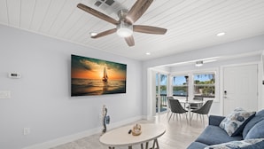 Una sala de estar moderna con ventilador de techo, un cuadro de un atardecer sobre el océano, un comedor con sillas y vista a través de la puerta de vidrio.