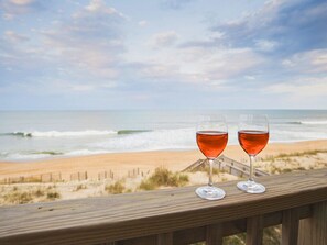 Two glasses of red wine on a wooden deck overlooking a beach with waves and a wooden fence. - Oceanfront 2BR/2BA Condo | Pet Friendly | Kill Devil Hills OBX (Kill Devil Hills)