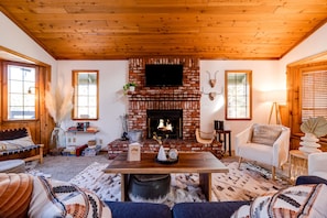 A cozy living room with a brick fireplace, wooden ceiling, and large windows.