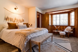 A bedroom with a large bed, a bench, a window with blinds, and a wall-mounted lamp.