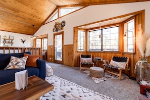 A cozy cabin interior with a wooden ceiling, large windows, a sofa, a chair, and a coffee table.
