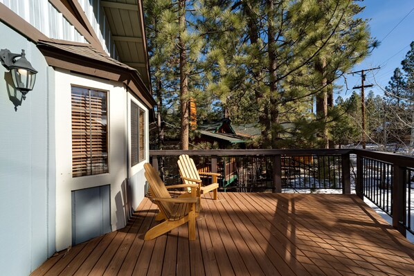 A wooden deck with two Adirondack chairs, a railing, and a view of tall pine trees.
