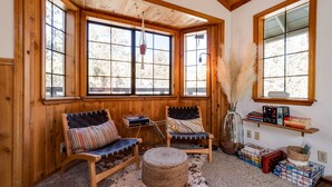 A cozy room with wooden walls and large windows, featuring two chairs, a rug, and a shelf with books and boxes. - Hot tub, bbq, 2 fireplaces, 5 mins to slopes & lake (Big Bear Lake)