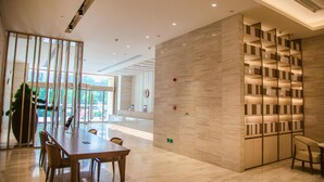 Un vestíbulo moderno de hotel con un amplio mostrador de recepción de madera, un área de descanso con sillas, una gran planta en maceta y una estantería montada en la pared.