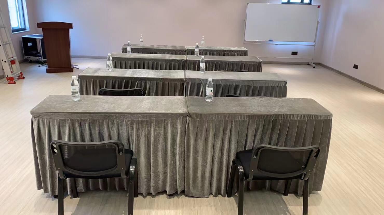 A conference room with four tables, each covered with a grey tablecloth and equipped with water bottles. There are two black chairs at the front table. A whiteboard and a podium are visible in the background.