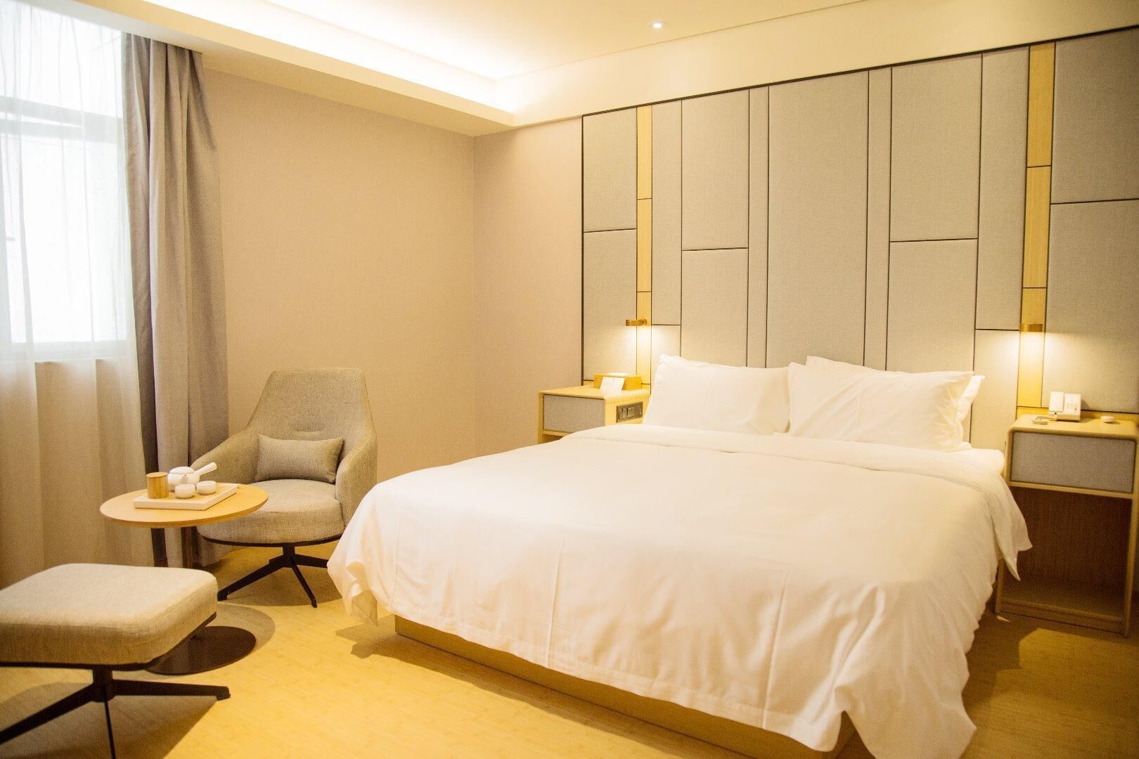 A hotel room with a large bed, a chair, a small table, and a nightstand with a lamp.