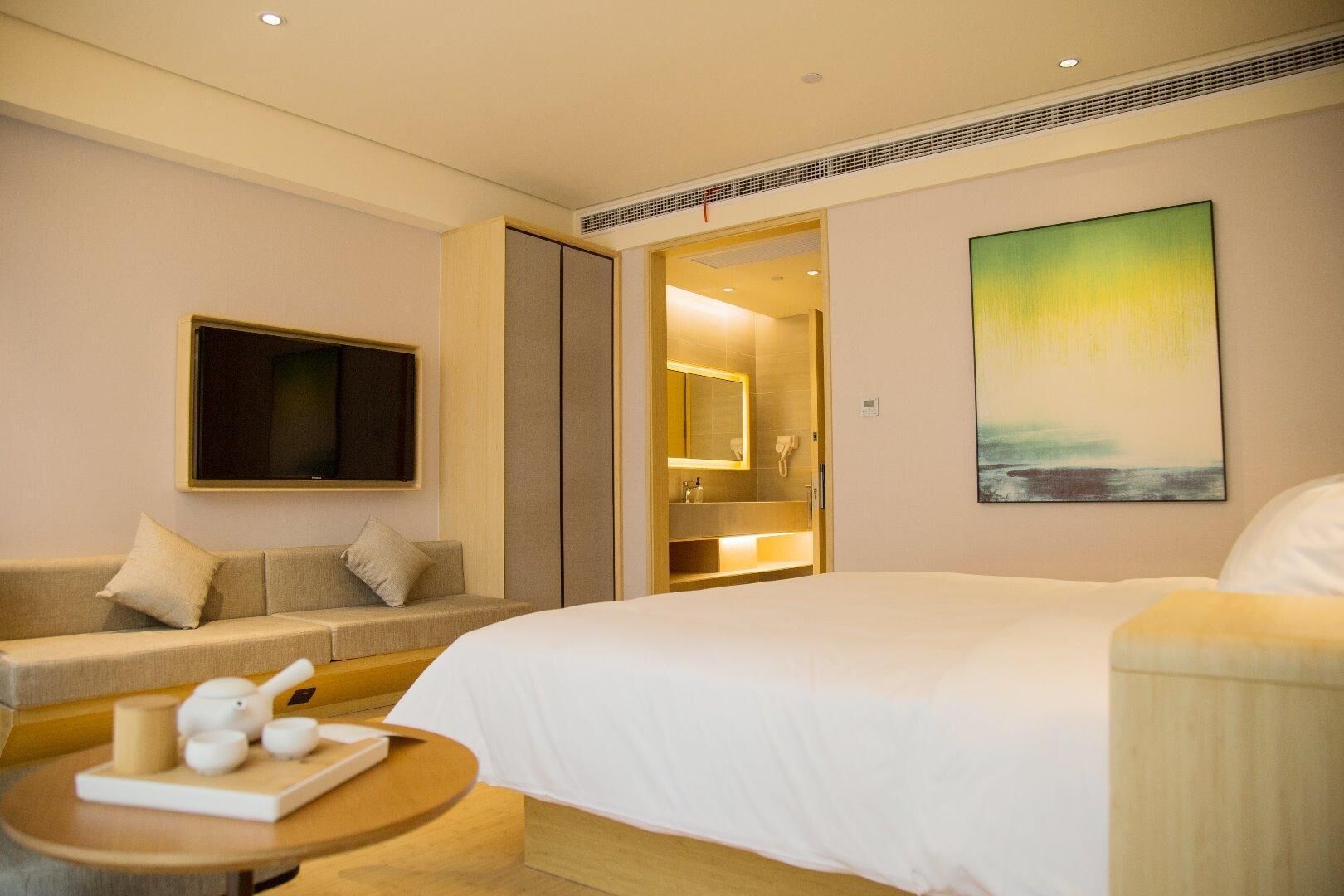 A hotel room with a bed, a sofa, a TV, a bathroom, and a painting on the wall.