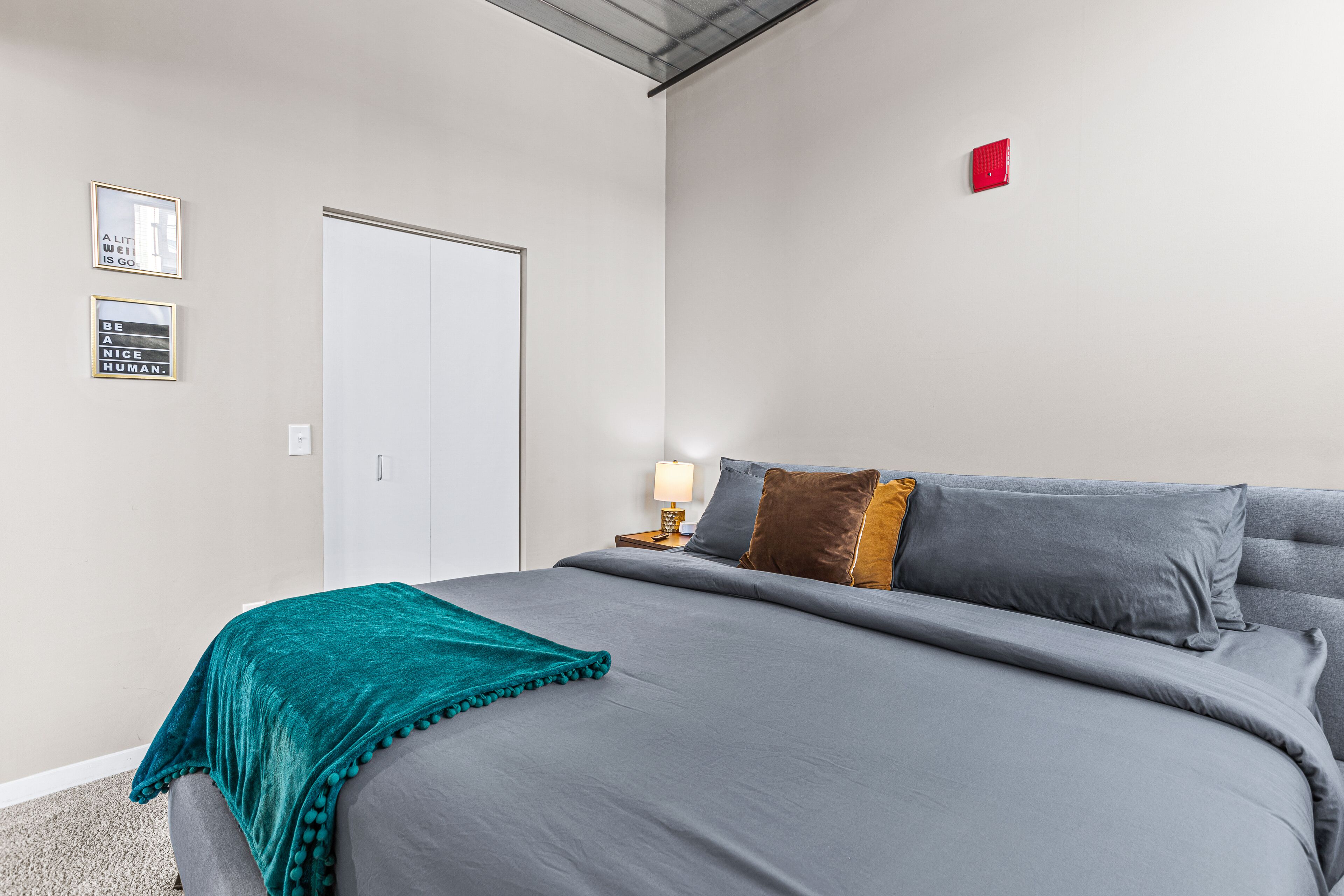 A neatly made bed with a teal throw blanket, a grey duvet, and multiple pillows. There is a nightstand with a lamp and a small table beside the bed. The room has a white door and a red light on the wall.
