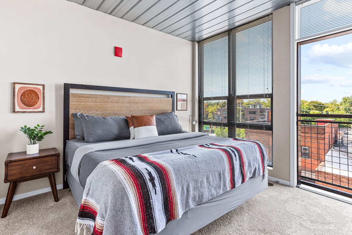 Un dormitorio moderno con una cama grande, una mesita de noche con una planta y vistas a la ciudad a través de amplias ventanas.