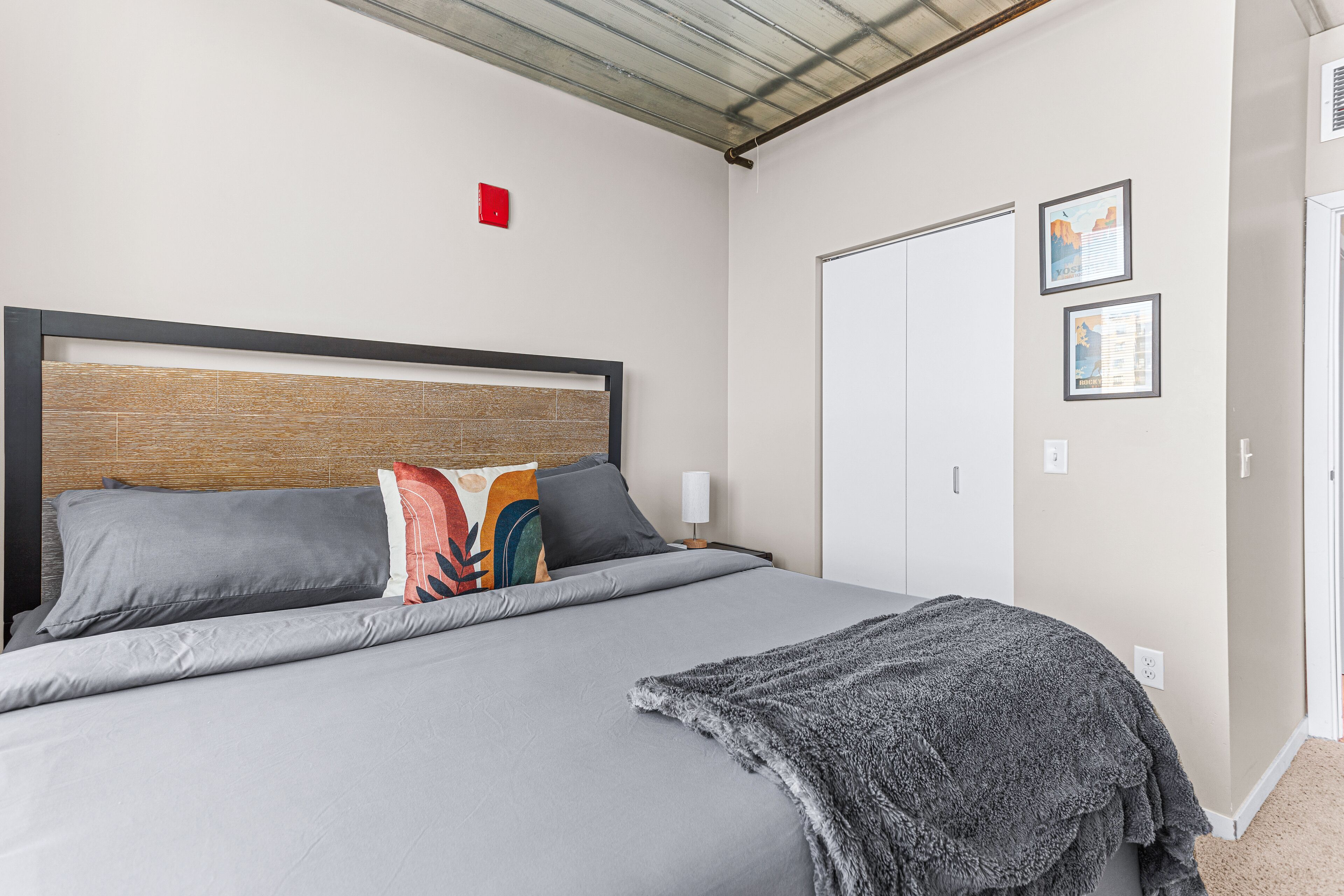 A neatly made bed with grey bedding and a grey throw blanket, a wooden headboard, and a white door in the background.