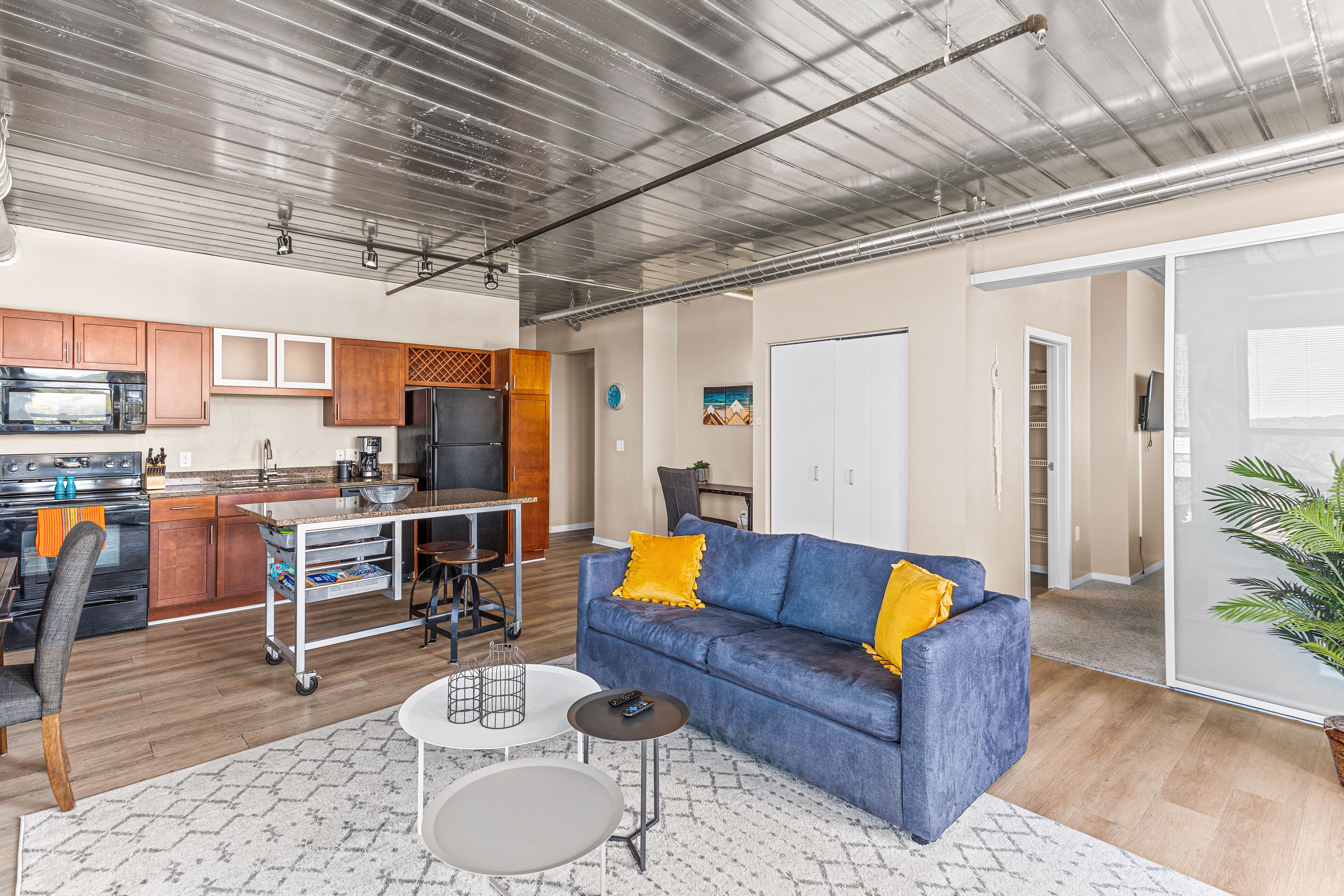A modern kitchen with stainless steel appliances, a dining area with a blue sofa and a small table, and a living room with a large window.