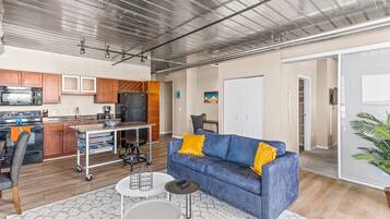 A modern kitchen with stainless steel appliances, a dining area with a blue sofa and a small table, and a living room with a large window.
