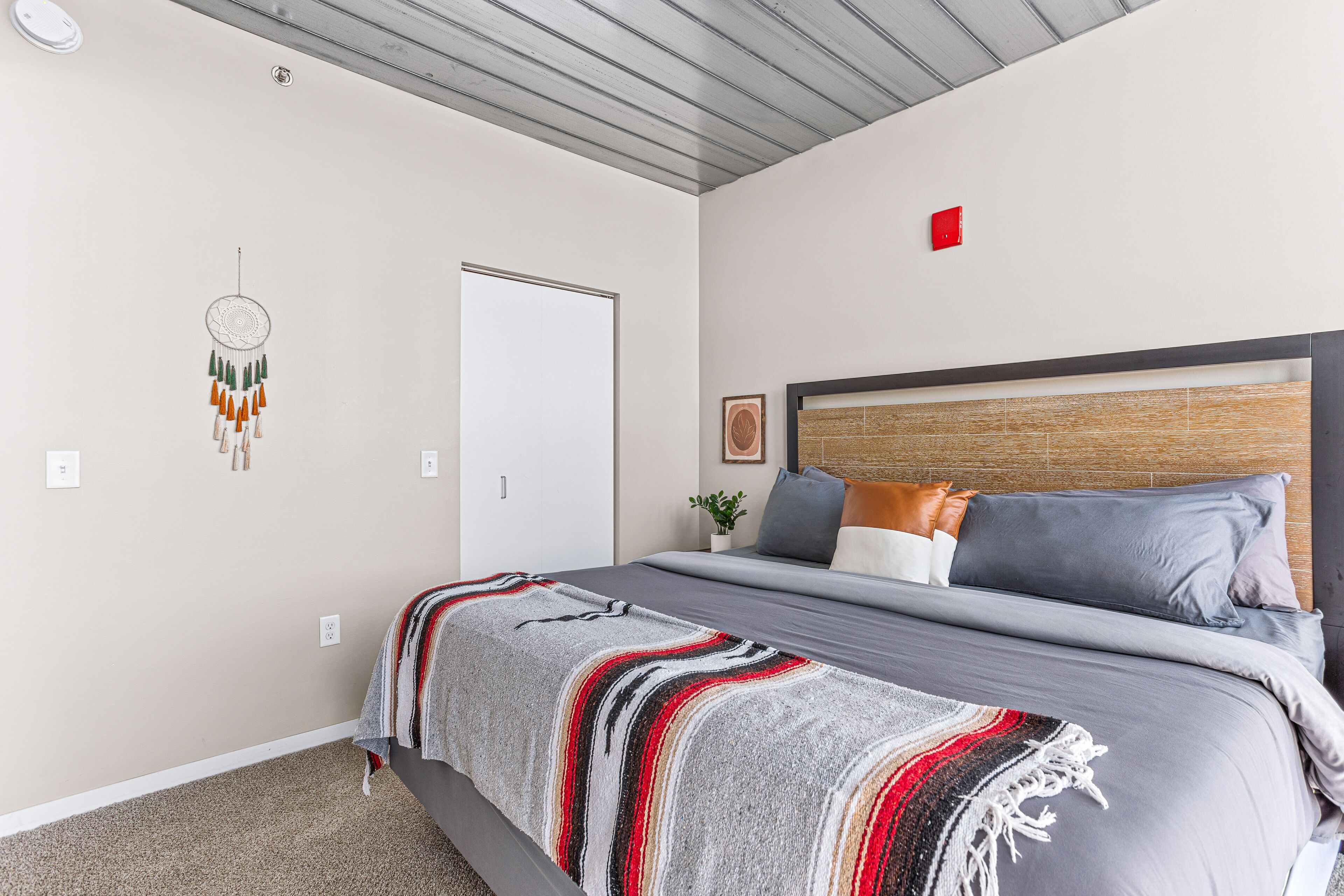 A bedroom with a bed, a headboard, a wall-mounted dreamcatcher, a door, and a red emergency light.