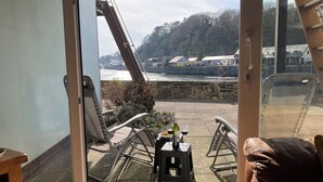 A view from a room with a large window overlooking a riverside area with outdoor seating and a stone wall. - 48A South Snowdon Wharf (Porthmadog)
