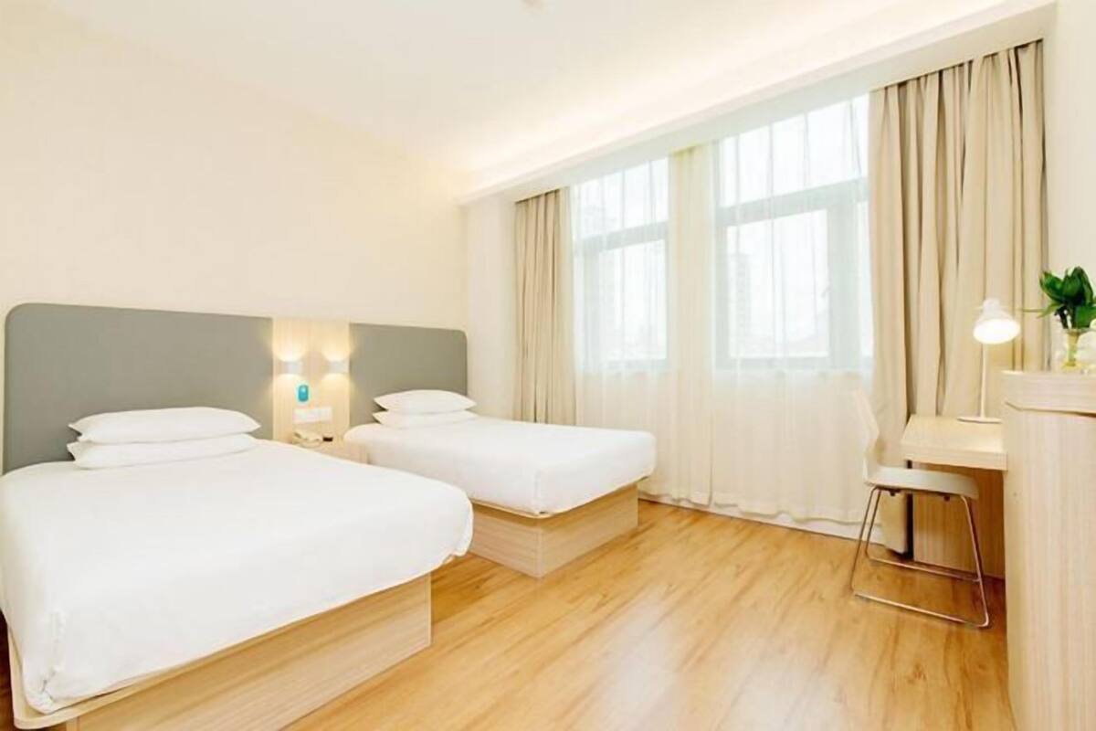 A hotel room with two single beds, a desk with a chair, and a large window with curtains.