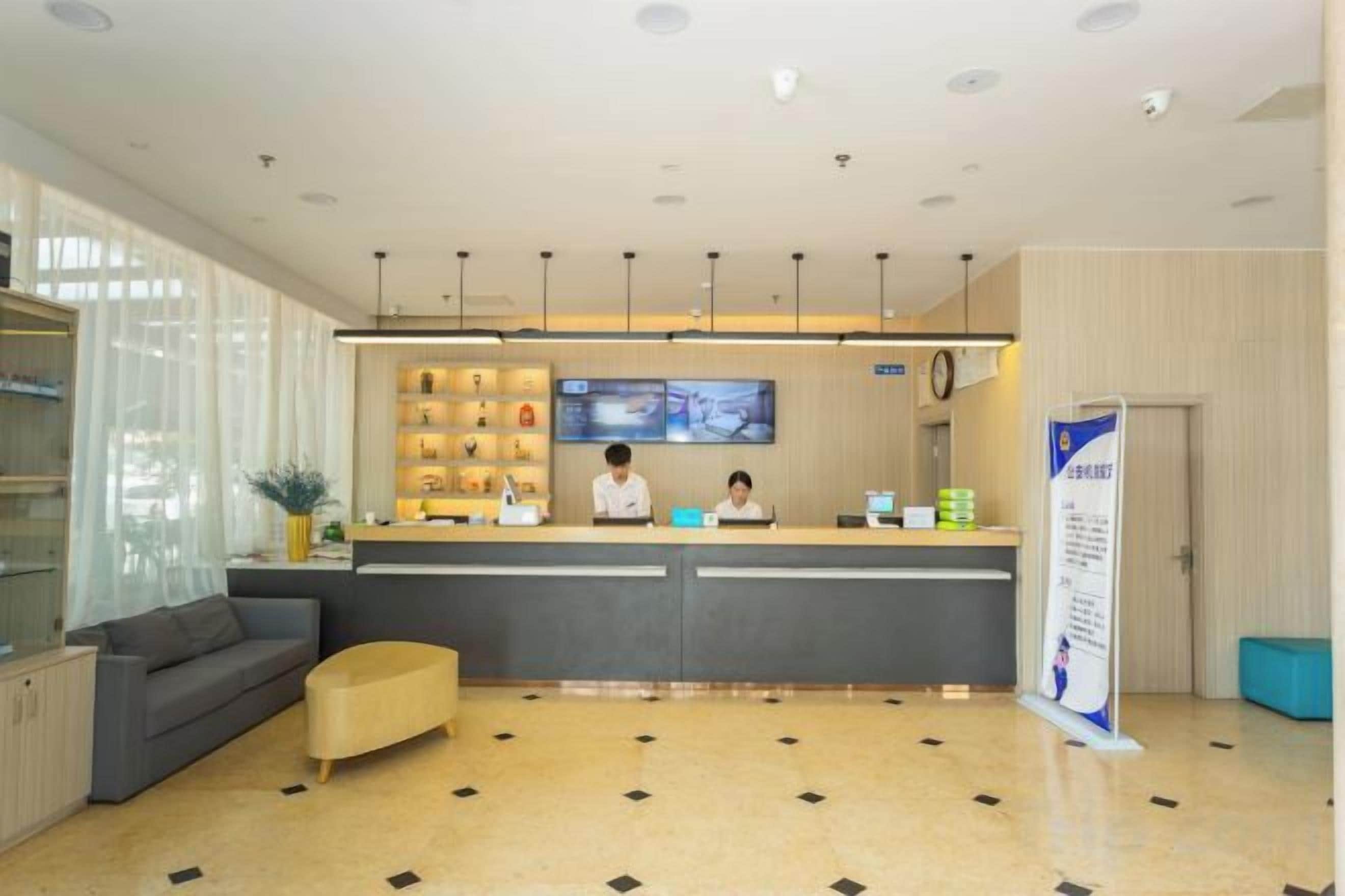A hotel reception area with a reception desk, a seating area with a sofa and ottoman, a television screen, and a wall-mounted shelf with items.