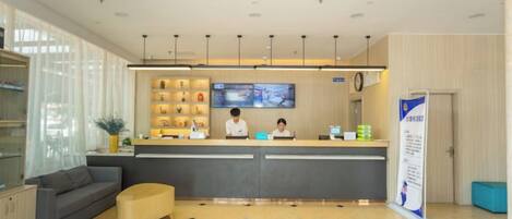 A hotel reception area with a reception desk, a seating area with a sofa and ottoman, a television screen, and a wall-mounted shelf with items.